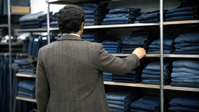 Man browsing stacks of folded jeans in a clothing store, representing denim fashion, retail shopping, apparel selection, and consumer lifestyle
