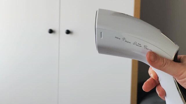 Compact electric garment steamer body profile in modern bedroom interior. White garment steamer showcases ergonomic handle, power specifications, and sleek design for convenient home fabric care.