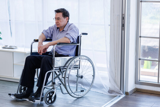 Sad elderly Asian man sitting in a wheelchair and looking down at his hand with a stressed looking thoughtfully near window,representing themes of health problems, aging society, depression in seniors