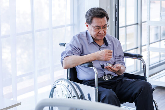 A middle-aged Asian man, a patient, sits alone in a wheelchair, holding tablets, pills, medications drugs, next to the window of his hospital room. There is space for a message.