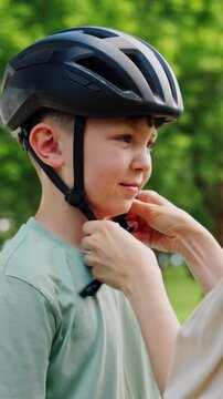Mother adjusts black helmet on boy before cycling in park