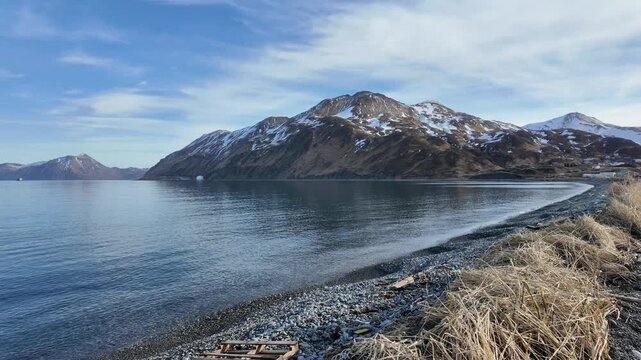 Scenic wide shot of dry beach grass and a pebble coast looking toward rugged snow dusted hills across a quiet bay.