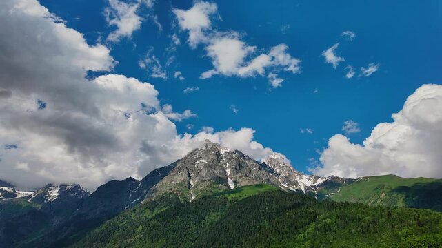 Majestic mountain range with snowy peaks and lush green forest on a sunny day with beautiful white clouds floating across the vibrant blue sky in a stunning natural landscape