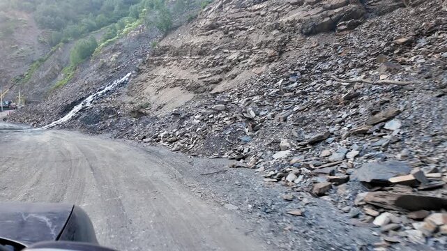 Personal perspective view from a vehicle cautiously driving on a perilous, unpaved mountain road next to a rocky slope with a stream of water flowing from a recent landslide