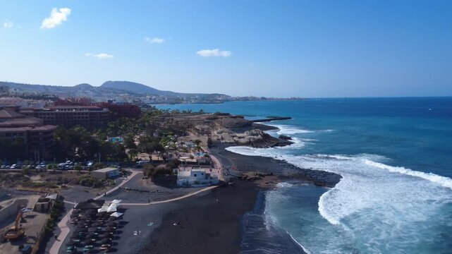 Vista a&eacute;rea de olas del Atl&aacute;ntico llegando a una playa volc&aacute;nica de arena negra con monta&ntilde;as al fondo, La Caleta, Costa Adeje, Tenerife