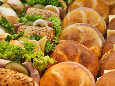 Rows of fresh ham and cheese bread rolls alongside garnished party sandwiches served on a catering tray for a breakfast or lunch buffet.
