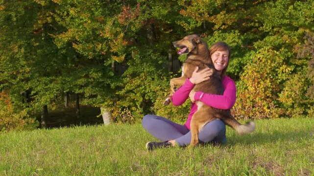 CLOSE UP, PORTRAIT: Cheerful woman sits in a sunny autumn meadow as her affectionate mixed breed dog comes to her for attention. Playful pet places its paws on her lap while she cuddles and pets it.