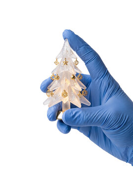 A hand in a blue medical nitrile glove holding a glowing decorative frosted glass Christmas tree ornament, transparent background