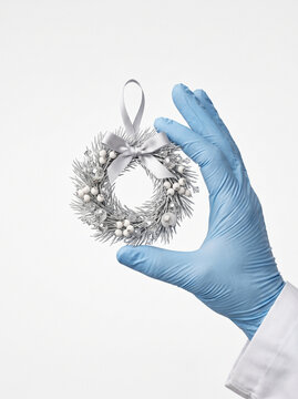 A hand in a blue medical glove holds a small silver Christmas wreath ornament against a white background