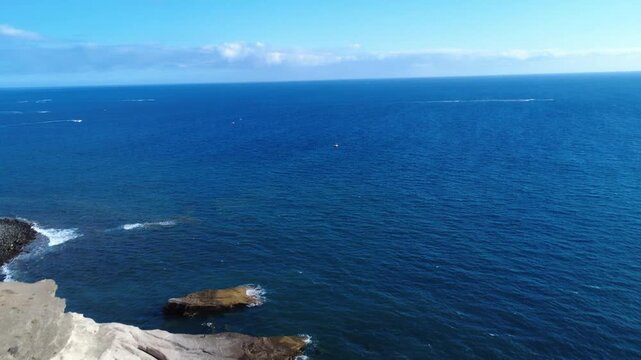 Dron vuela junto a acantilados volc&aacute;nicos claros y aguas atl&aacute;nticas azul profundo con un rompeolas y hoteles al fondo, La Caleta, Costa Adeje, Tenerife