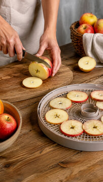 Person slicing fresh red apples on wooden cutting board to prepare them for food dehydration on round plastic mesh tray in kitchen setting.