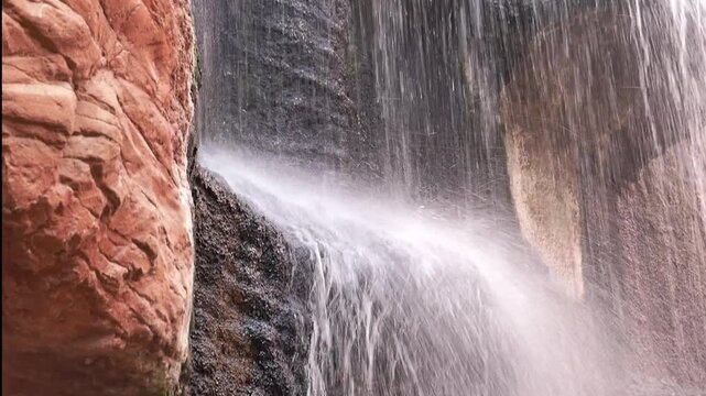 A red rock waterfall cascades over a sandstone cliff, creating misty spray and silky flowing water against textured rock layers in a quiet canyon setting, evoking nature, motion, and erosion