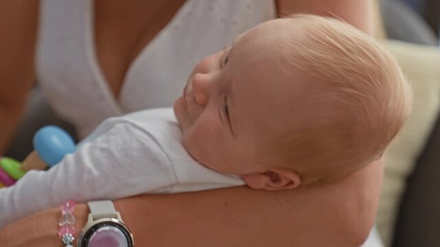 Blonde newborn baby cradled in mother's arms wearing a white onesie holding a colorful ring stacker rattle inside a building living room with soft sunlight; tenderness curiosity.