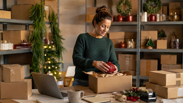 Woman working in warehouse packing red items into cardboard boxes surrounded by shelves and holiday decorations.