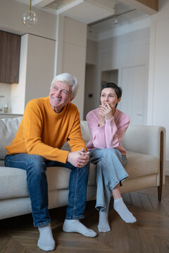 Happy senior married couple sitting together on sofa, looking away and smiling, elderly husband and wife watch television, talk and enjoy relaxed leisure time at home during retirement.