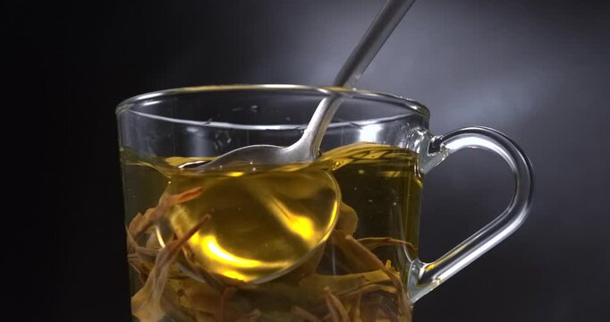 Hot green tea stirring with spoon in glass cup with steam on black background in slow motion