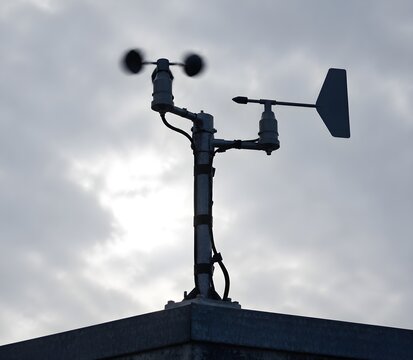 Anemometer and wind vane mounted on a rooftop mast against an overcast sky with spinning cups blurred by strong wind