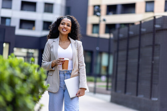 Smiling professional woman walking outdoors with takeaway coffee and laptop, representing commuting, career mobility and a modern urban work lifestyle