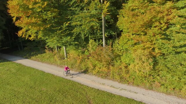 AERIAL: Elderly woman cycling an ebike along sunny gravel country road. A dense forest with green, yellow and orange foliage creates a colorful autumn backdrop beside an open meadow in rural Slovenia.