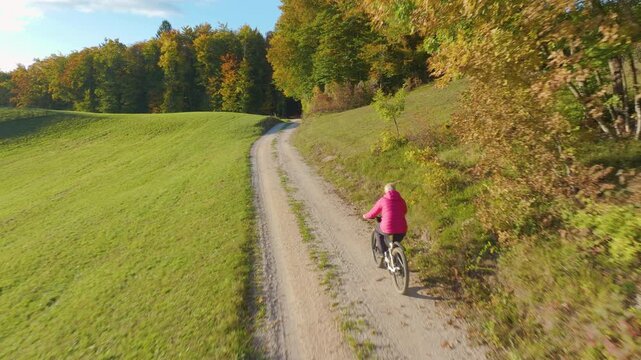 AERIAL: Senior lady riding an electric bike uphill on a gravel lane through rolling countryside. Sunny meadows and colourful autumn woodland surround the elderly cyclist in a peaceful rural Dolenjska.