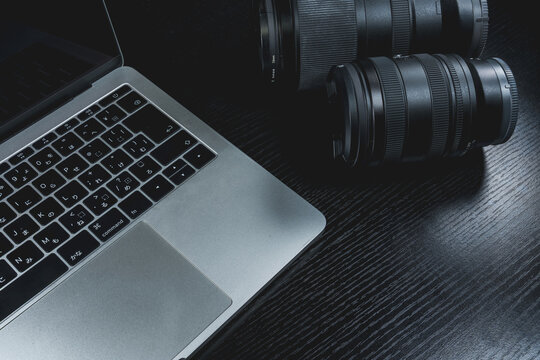 Close-up of two camera lenses sitting next to a laptop computer on a black wooden desk in dark tone. Perfect for professional photography, video editing, creative workspace, and tech backgrounds.