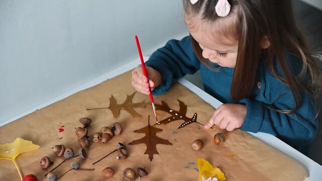 A little girl sits at a table and paints decorative patterns on dry autumn oak leaves with a brush. Creative craft lesson at home. Slow motion FHD footage