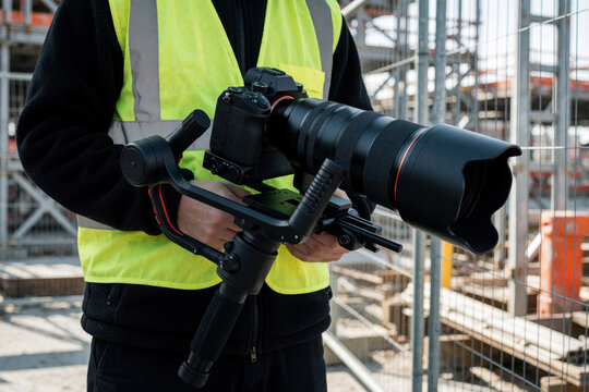 Pro videographer uses digital camera with large telephoto lens, electronic steadycam gimbal at construction site. Man wearing yellow safety vest holds heavy filming gear for media production. High