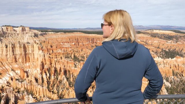 Blonde woman tourist wearing sunglasses and hoodie standing at overlook, contemplating stunning rock formations bryce canyon amphitheater on viewpoint in utah