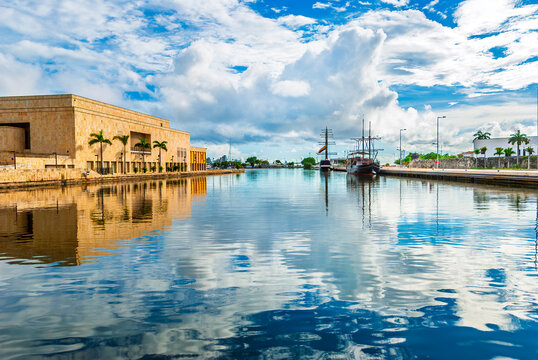 Cartagena port in Colombia with calm water reflecting a cloudy sky, a stone building, and vintage galleons docked in the Caribbean Sea.