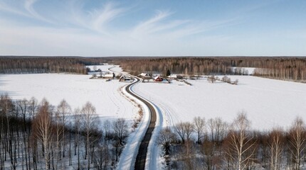 Fototapeta premium Aerial view of peaceful winter landscape. Winding asphalt road leads through vast snow covered fields toward remote village houses. Bare trees, small farm buildings sit in quiet countryside under