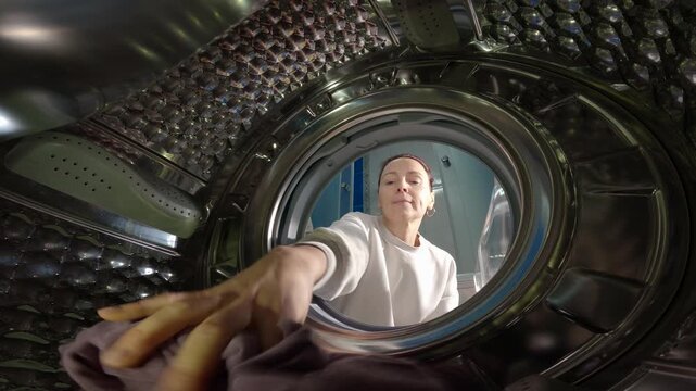 Woman loading dirty laundry into a washing machine. Unique perspective from inside a washing machine as a woman opens the door and puts colorful dirty clothes into the stainless steel drum