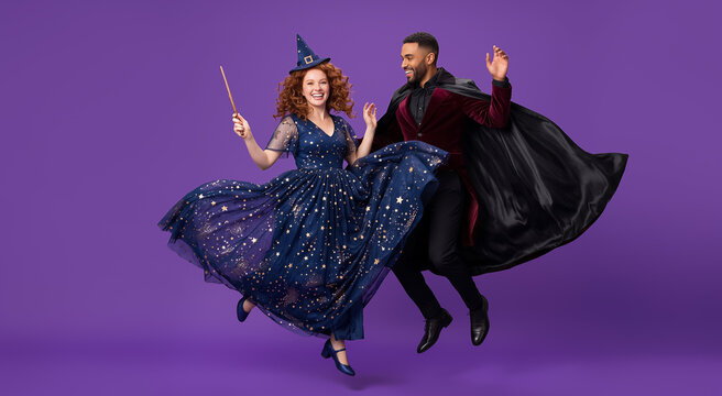 Woman in witch costume and man in wizard cape jumping joyfully against purple background. Magic fantasy concept, celebration, Halloween, cosplay.