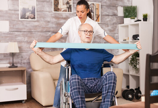 From the front an older wheelchair user stretches a resistance band while a therapist supports the exercise from behind during guided home rehabilitation and mobility training supporting recovery.