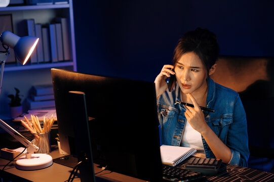 Young beautiful Asian creative woman calling on phone desk to customer or coworker explaining creator project's high profit while searching market analysis on pc at neon office at night. Stratagem.