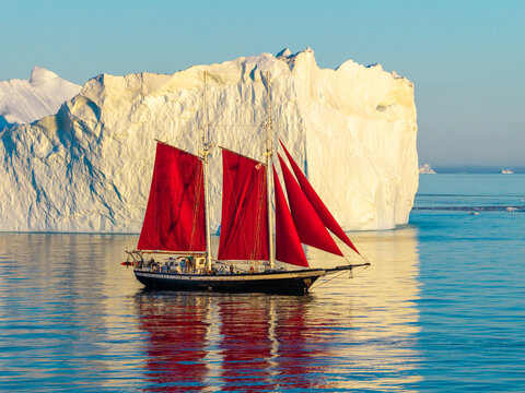 Aerial view of a sailboat with striking red sails navigating calm waters in front of a massive white iceberg in Greenland.