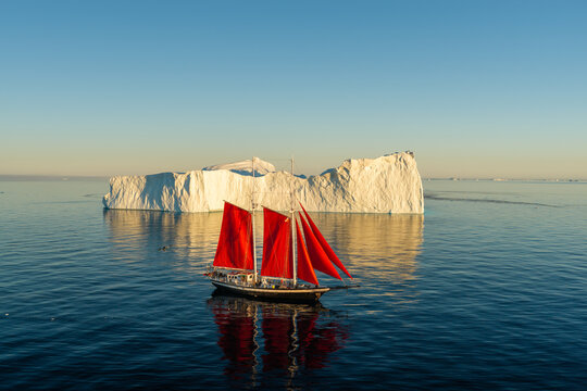 Aerial view of a sailboat with striking red sails navigating calm blue waters in front of a massive white iceberg under a clear sky in Greenland.