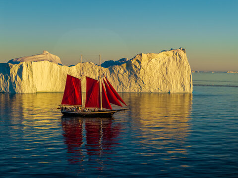 Aerial view of a sailboat with striking red sails gliding through calm blue waters past a massive, sunlit iceberg in the Arctic landscape of Greenland.