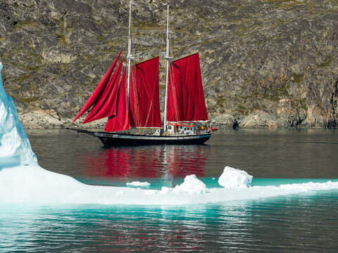 Aerial view of a sailboat with striking red sails navigating through calm icy waters past a large iceberg with a rugged rocky coastline in the background in Greenland.