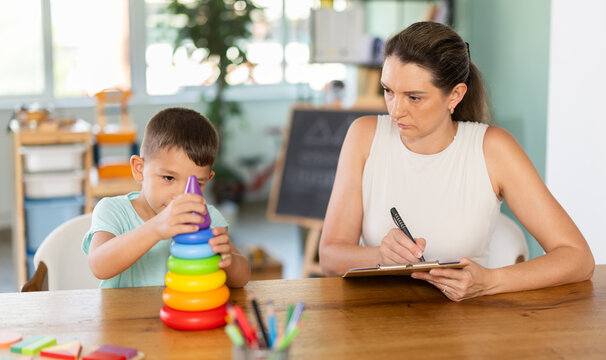 Professional child psychologist observing little boy assembling toy pyramid on table at development center