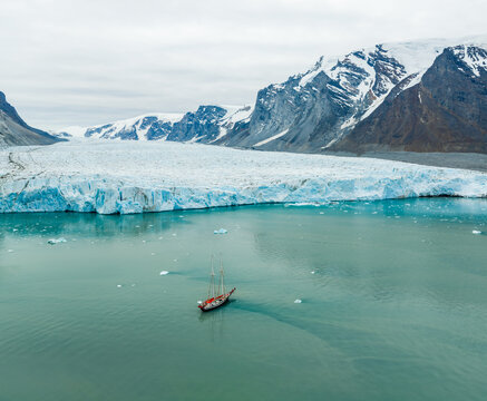 Aerial view of a sailboat navigating the turquoise waters of a glacial bay, with a massive glacier wall and snow-capped mountains in the background in Greenland.