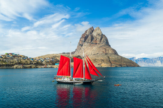 Aerial view of the Uummannaq mountain rising above the sea with a sailboat featuring striking red sails navigating the blue waters near the colorful houses of Uummannaq, Kitaa, Greenland.