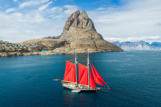 Aerial view of a sailboat with striking red sails navigating the blue waters near the iconic Uummannaq mountain and the colorful coastal town of Uummannaq, Kitaa, Greenland.