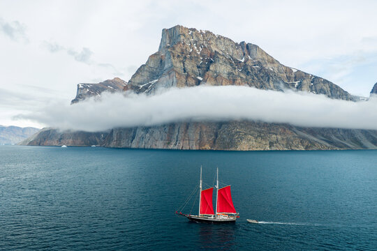 Aerial view of a sailboat with red sails in a fjord, set against the dramatic backdrop of Storoen mountain partially shrouded in low-hanging clouds in Avannaata Kommunia, Greenland.