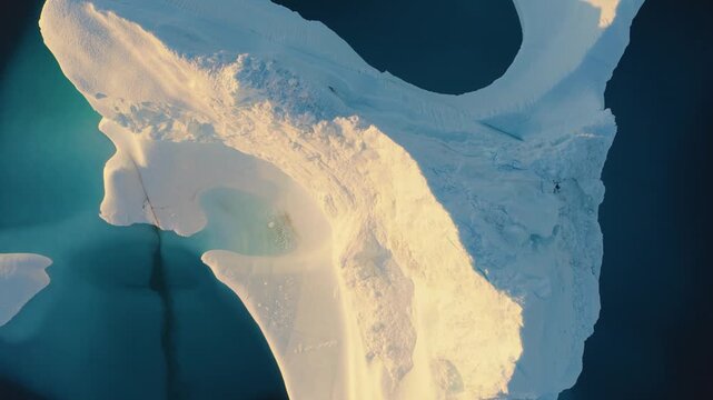 Aerial view of a massive iceberg floating in the deep blue ocean, showcasing intricate ice textures and golden sunlight reflecting off the frozen surface in Greenland.