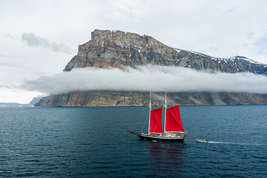 Aerial view of a sailboat with striking red sails navigating the deep blue waters near the dramatic cliffs of Storoen, Avannaata Kommunia, Greenland.