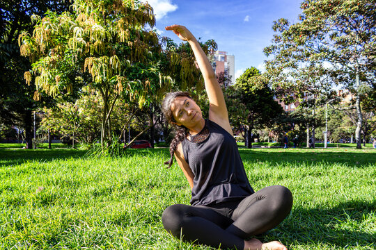 Young woman sitting cross-legged on the grass and doing side stretch exercises in a sunny public park, practicing outdoor yoga and flexibility routine.