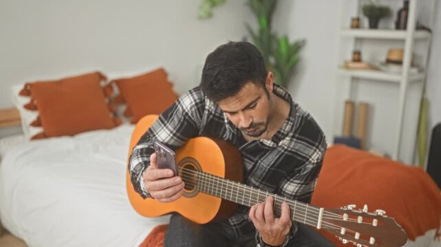 Man playing classical guitar, left hand on fretboard and right hand strumming while holding smartphone in bedroom; quiet concentration.