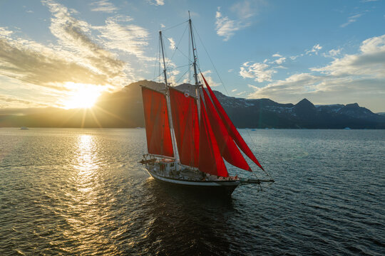 Aerial view of a sailboat with striking red sails navigating calm waters during a golden sunset with mountainous terrain in the background, Greenland.