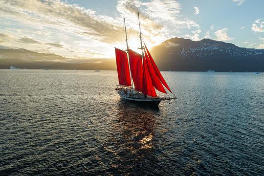 Aerial view of a sailboat with striking red sails navigating calm waters during the golden hour, with distant mountains and icebergs under a soft sky in Greenland.
