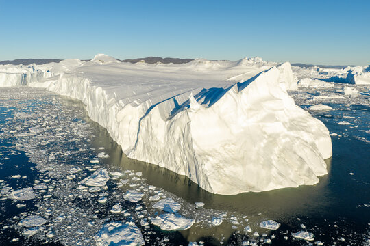 Aerial view of the massive Ilulissat glacier iceberg floating in dark blue arctic waters with scattered ice fragments under a clear sky in Greenland.